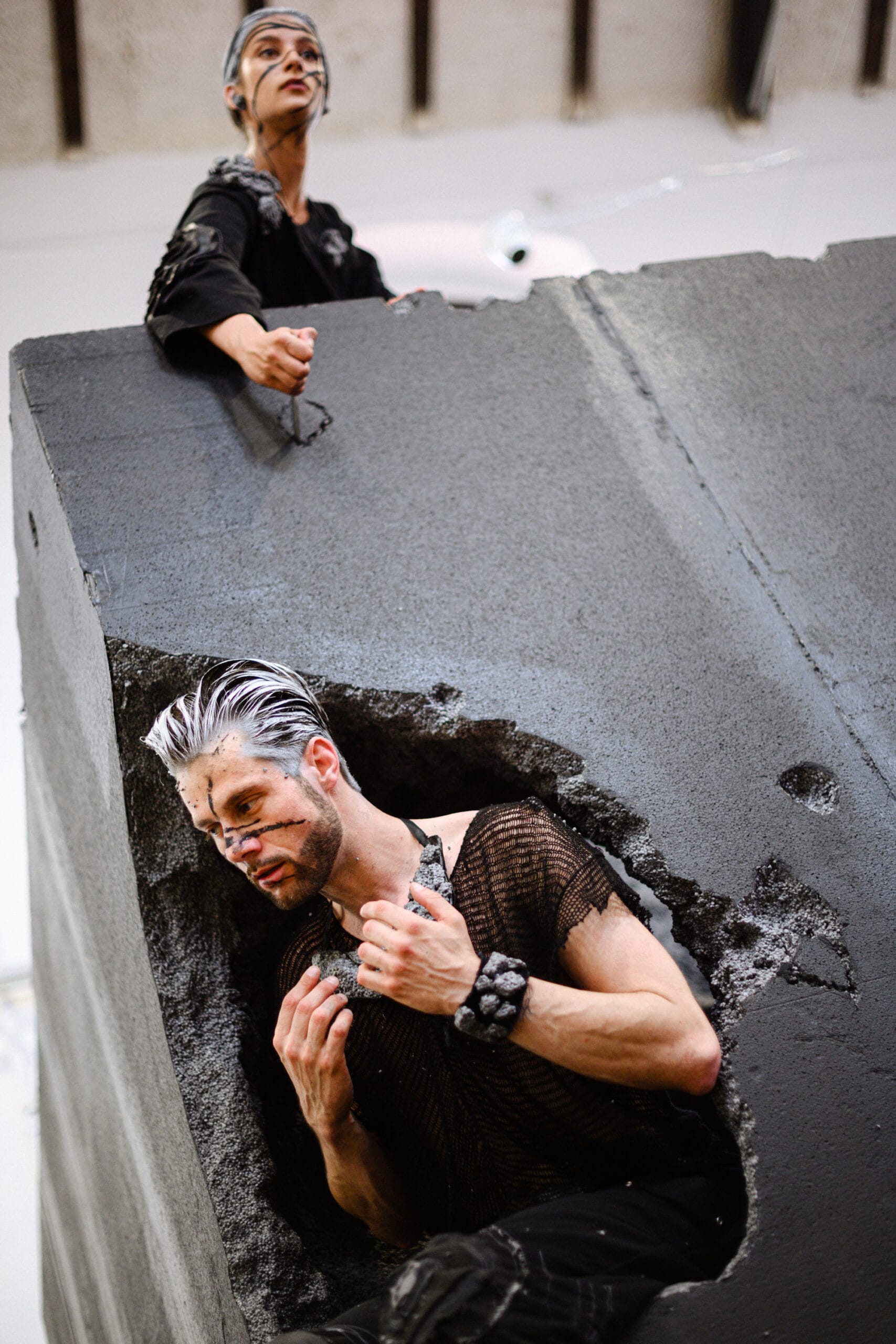 Two performers in dark costumes emerge from a broken concrete stage set, one climbing through a jagged opening.