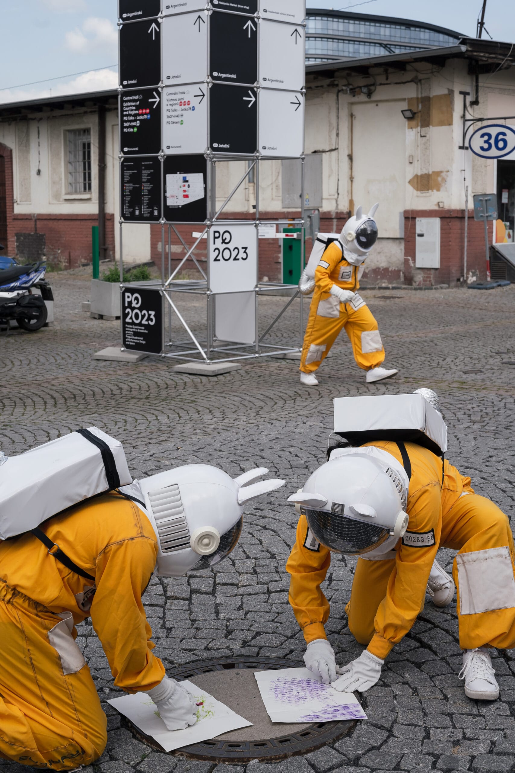 Performers in yellow spacesuit costumes crouch on cobblestones near a PQ 2023 signage tower in Prague.
